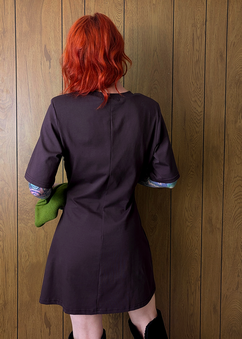 Person wearing a dark plum t-shirt dress against a wooden wall.