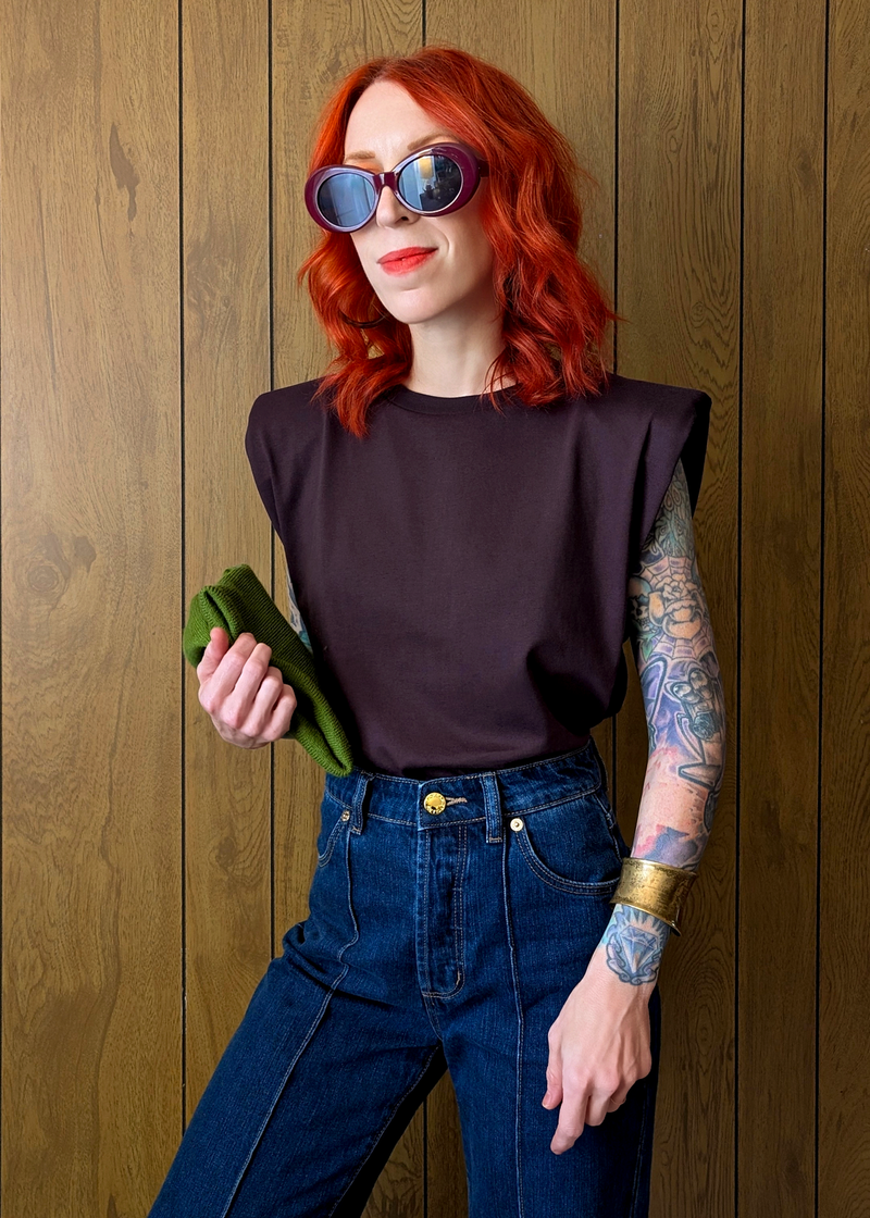Person with red hair wearing sunglasses, a dark plum top, and blue jeans against a wooden wall.