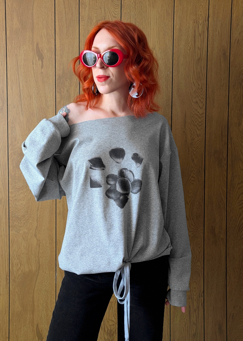 Person wearing a heather grey off shoulder jersey top with black floral graphics, black pants, and red sunglasses against a wooden wall.