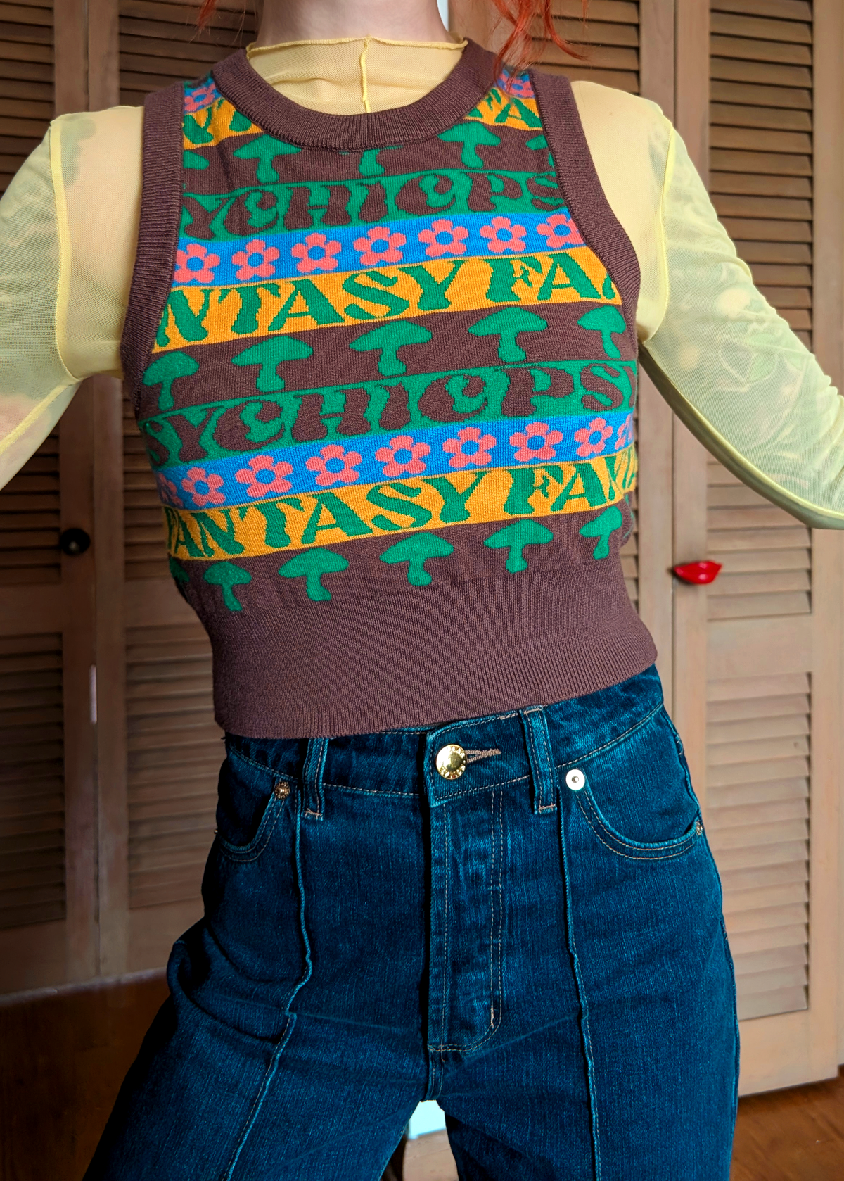 Person wearing a colorful patterned sweater vest over a light yellow mesh shirt with blue jeans.