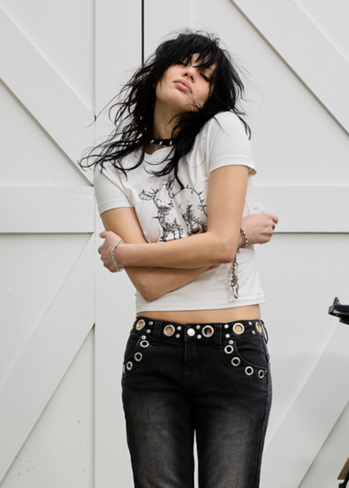 Person wearing an ivory graphic t-shirt  with spiked lamb design, and black pants with grommets against a white background