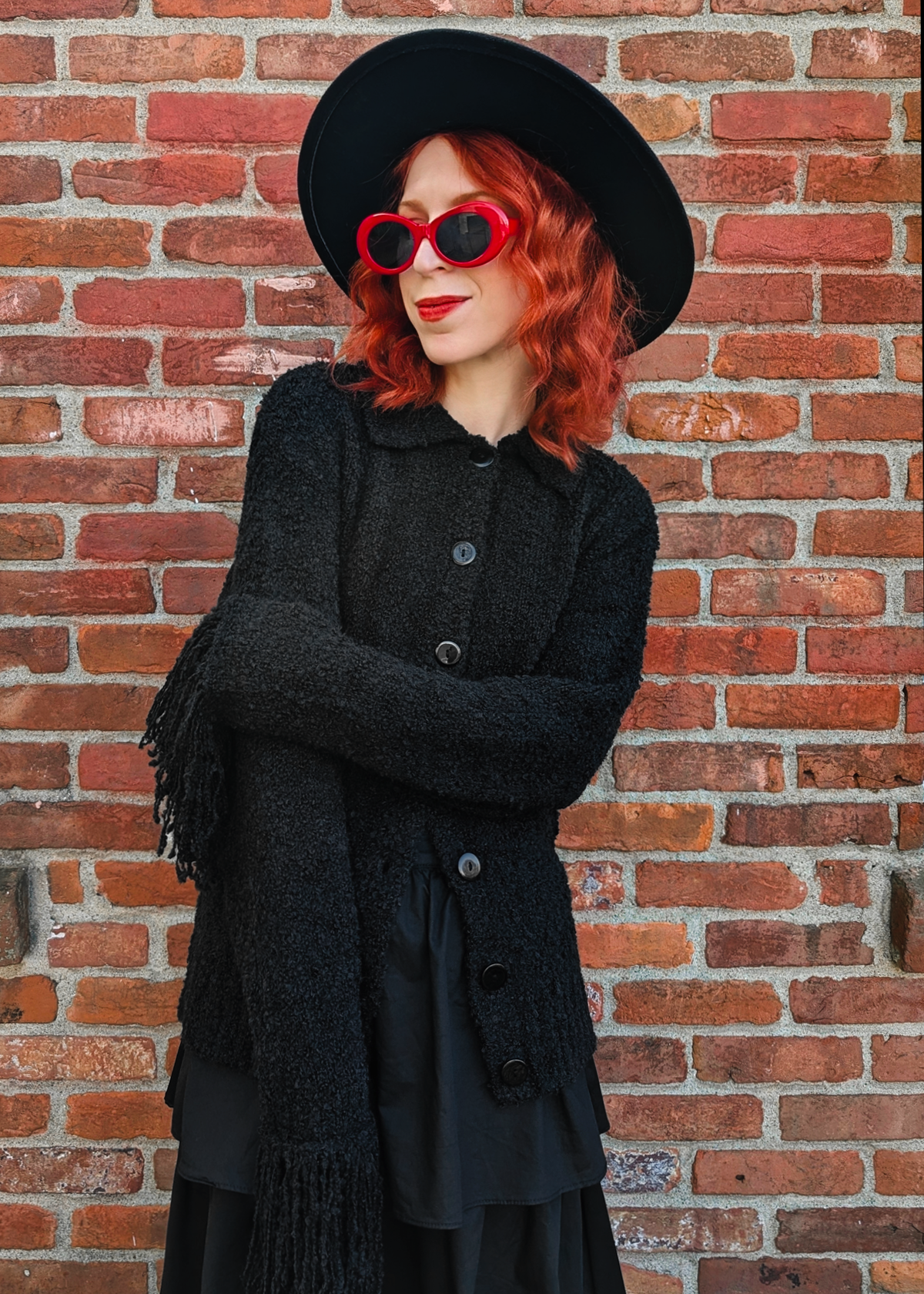 Person with red hair wearing a black fuzzy cardigan with fringe sleeves, a black hat, and sunglasses against a brick wall.