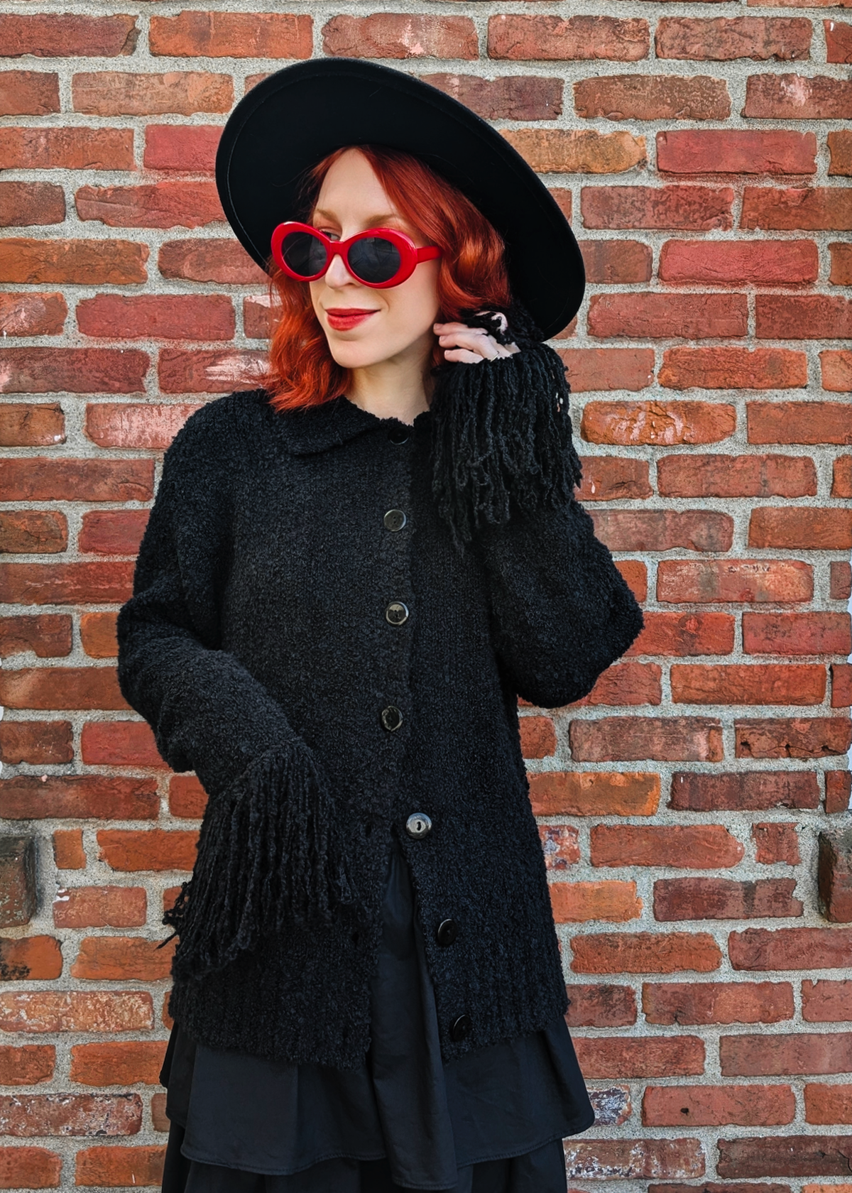 Person wearing a black cardigan, hat, and sunglasses against a brick wall.