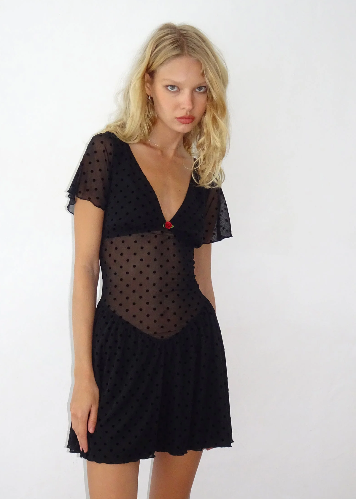 90s inspired black mesh mini dress with black velvet flocked dots allover. Features flutter short sleeves, v-neckline with red satin rosette, sheer waist, and floaty skirt. By Motel Rocks