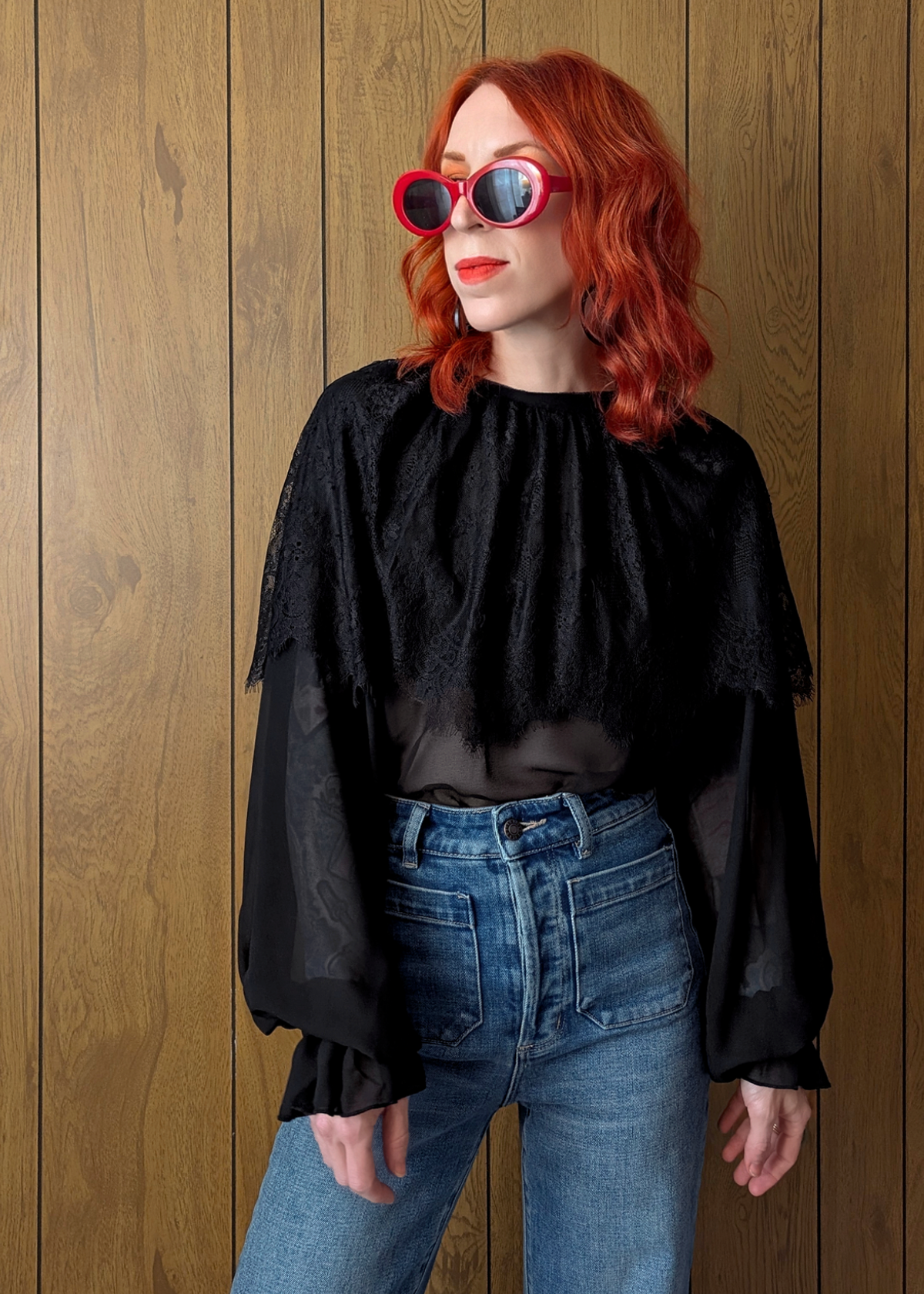 Person wearing a black lace chiffon top and blue jeans with red sunglasses against a wooden panel background