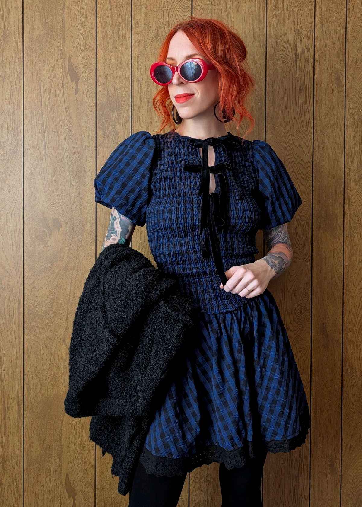 Person wearing a navy and black checkered dress with a black sweater on their arm against a wooden background