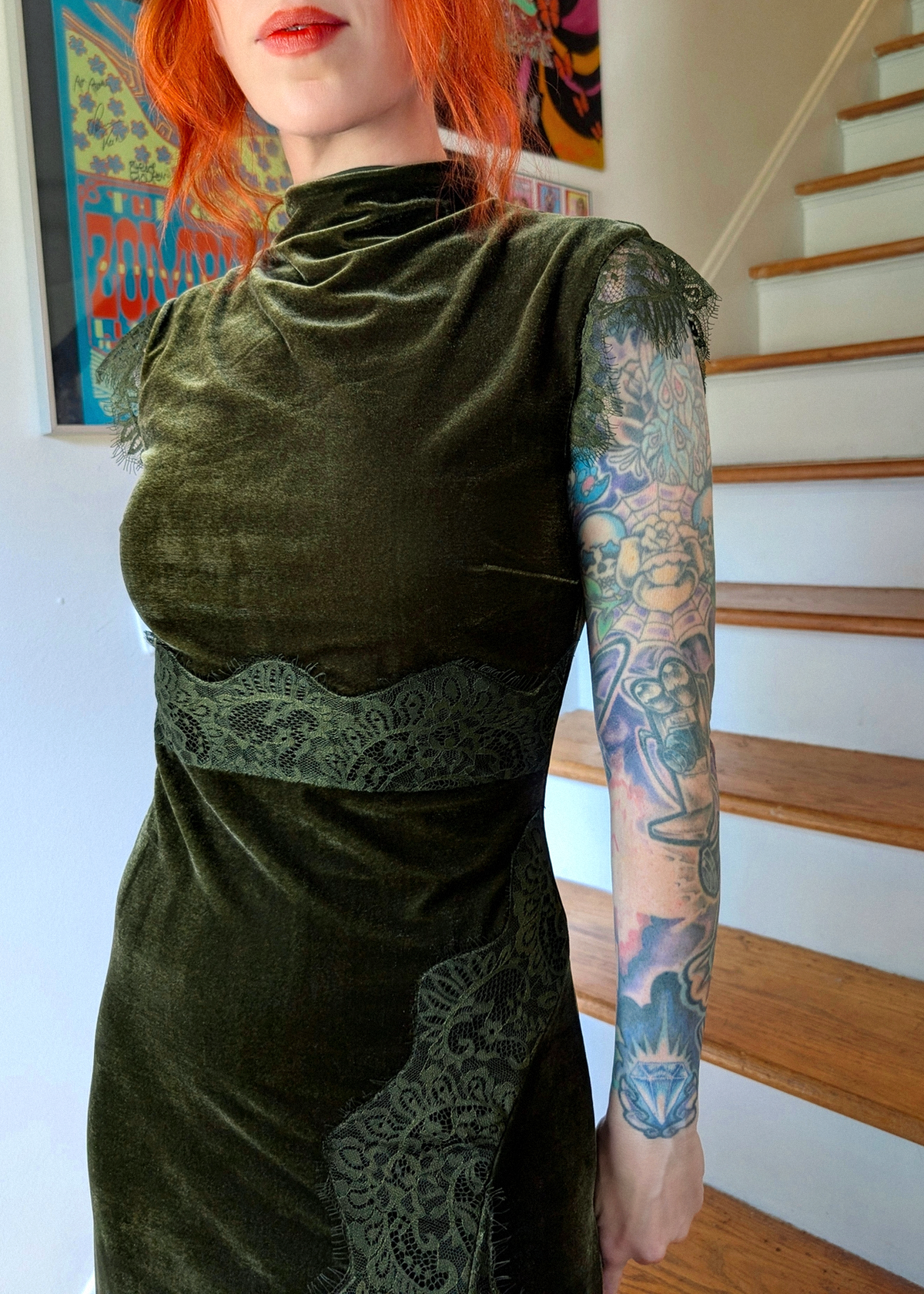 A woman with bright red hair and a tattooed arm wearing a dark olive green velvet dress with lace trim, standing in front of a staircase.