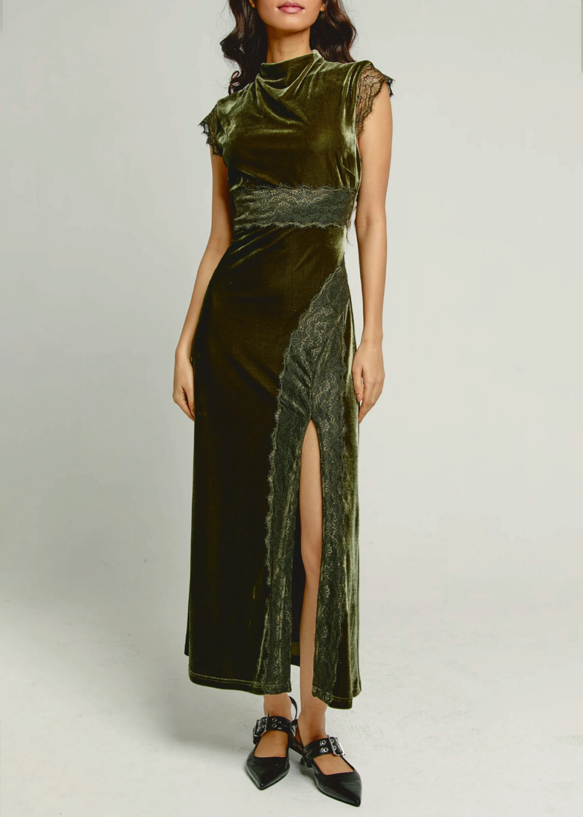 A woman models a maxi-length olive green velvet gown with sheer lace cap sleeves, a matching lace waistband, and a high thigh slit. She is wearing black pointed-toe flats against a plain light gray studio background.