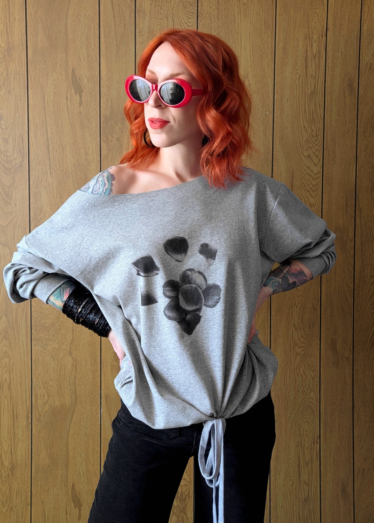 Person wearing a heather grey off shoulder jersey top with black floral graphics, black pants, and red sunglasses against a wooden wall.