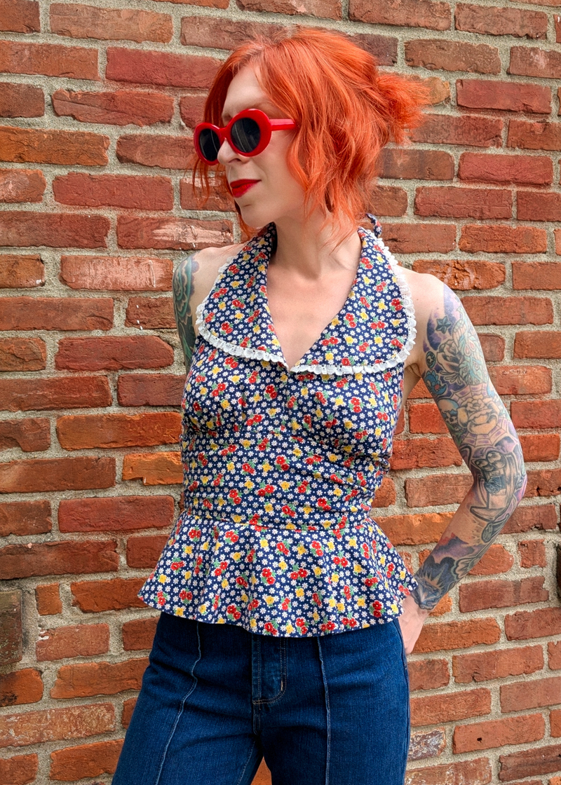 Person with red hair and tattoos wearing a vintage floral halter top with eyelet trim collar standing against a brick wall background