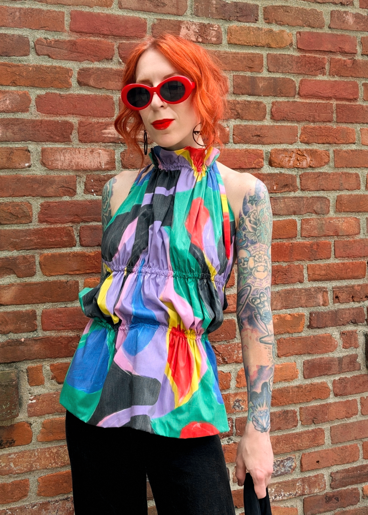Person with colorful ruched halter top and sunglasses standing against a brick wall