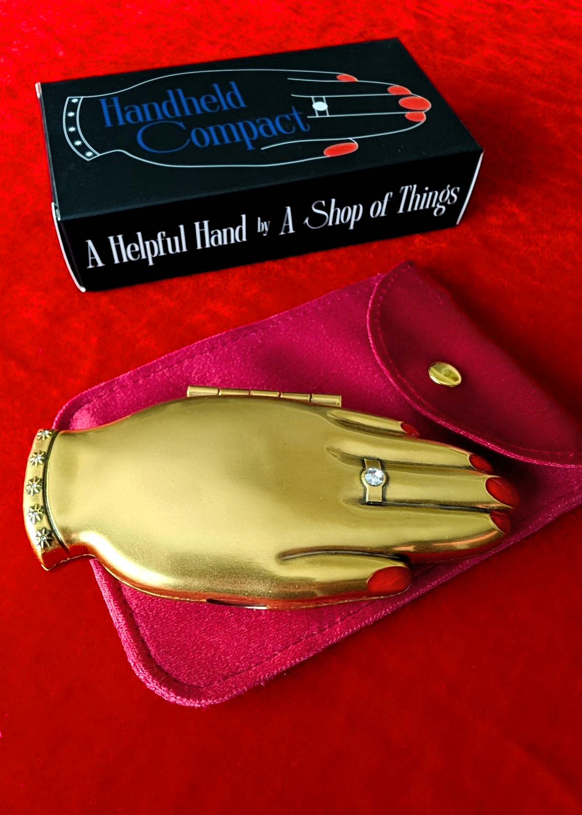 A gold hand-shaped compact mirror displayed on a magenta velvet pouch, with its black retail box visible behind it on a red fabric background. The box reads 'Handheld Compact' and 'A Helpful Hand by A Shop of Things.