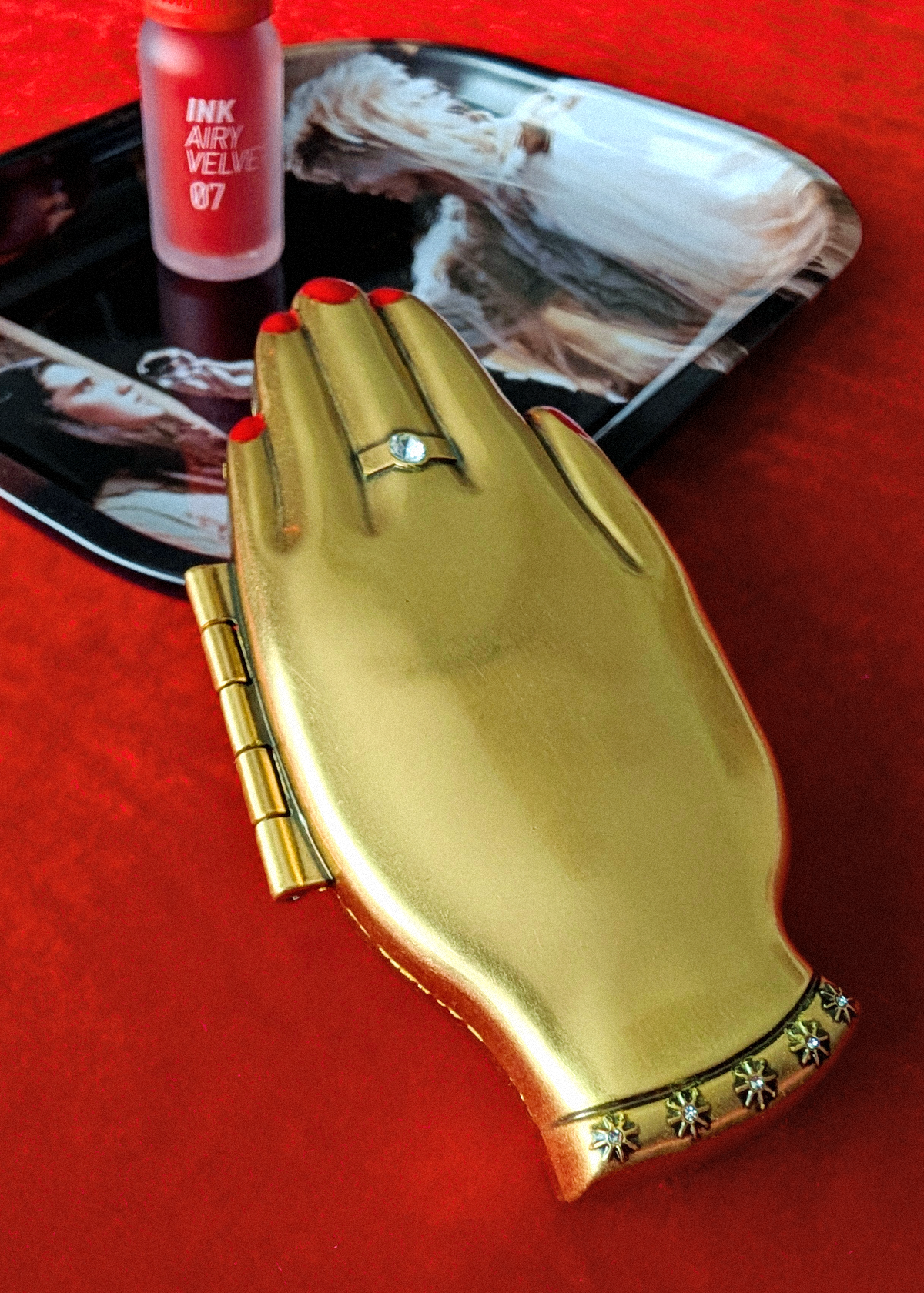 A gold hand-shaped compact case with a hinge closure and engraved embellishments on the wrist, displayed on a red textured surface with a tray and lipstick in the background