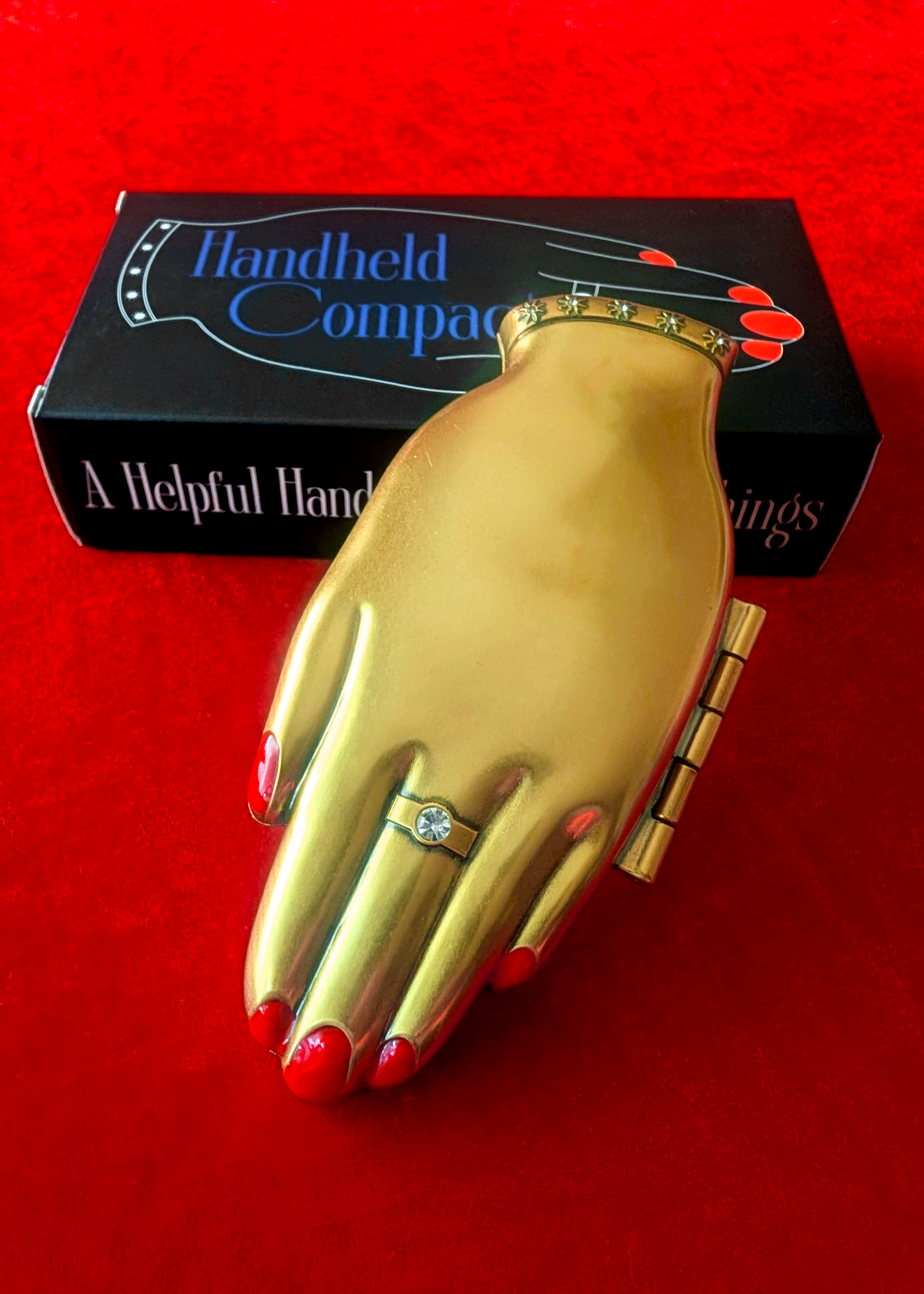 A gold hand-shaped compact with red painted fingernails and a ring on the middle finger, displayed in front of its black packaging box on a red velvet background.