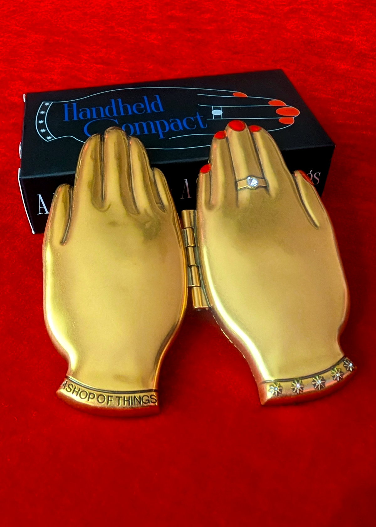A gold hand-shaped compact case opened up showing both sides of the compact, placed in front of its black branded box on a red textured background. The left wrist area reads 'A SHOP OF THINGS' and the right wrist has a ring with red nail polish detail.
