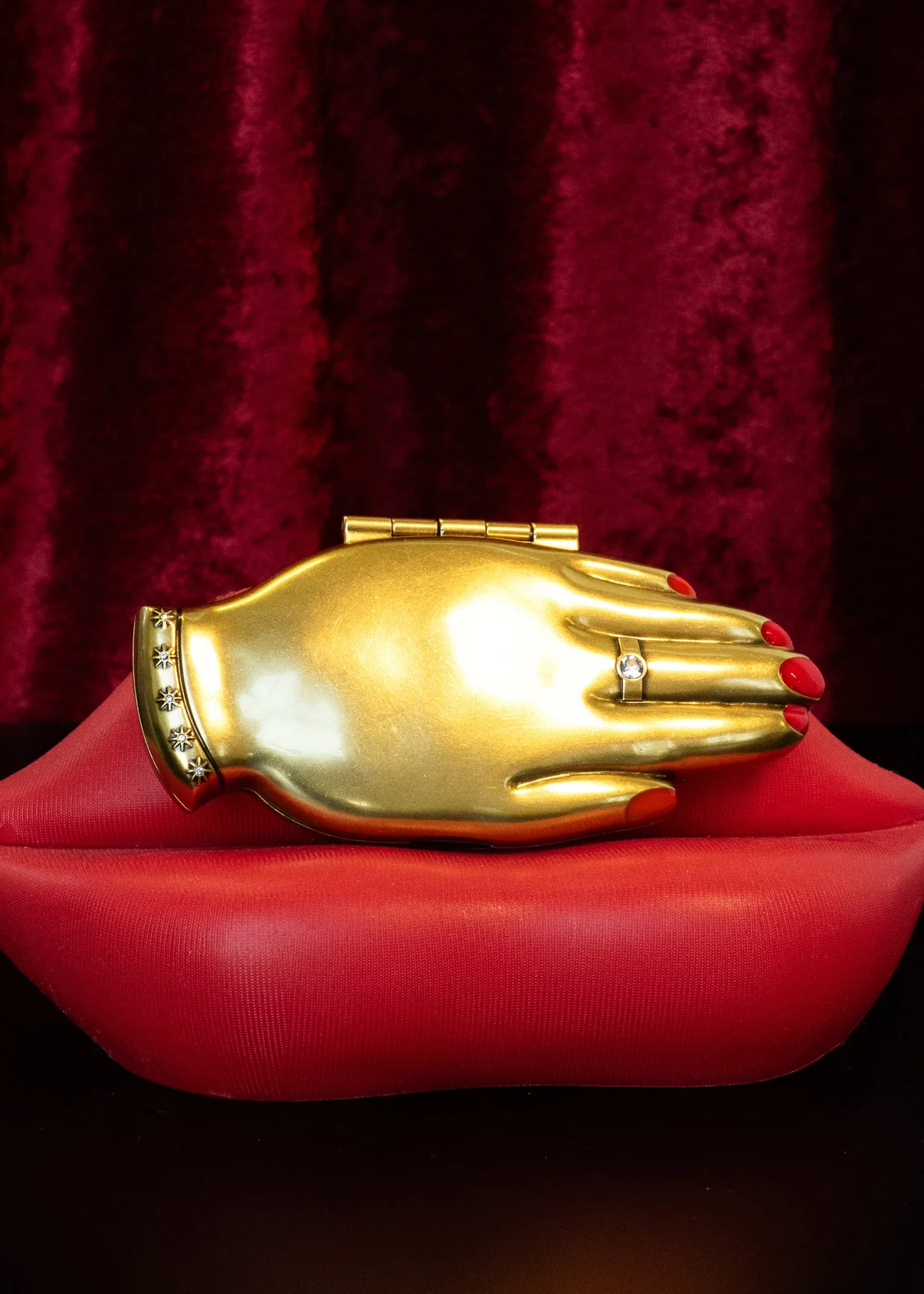 A gold hand-shaped compact mirror with red nail details and a ring, resting on a red lips-shaped base against a dark red velvet background.