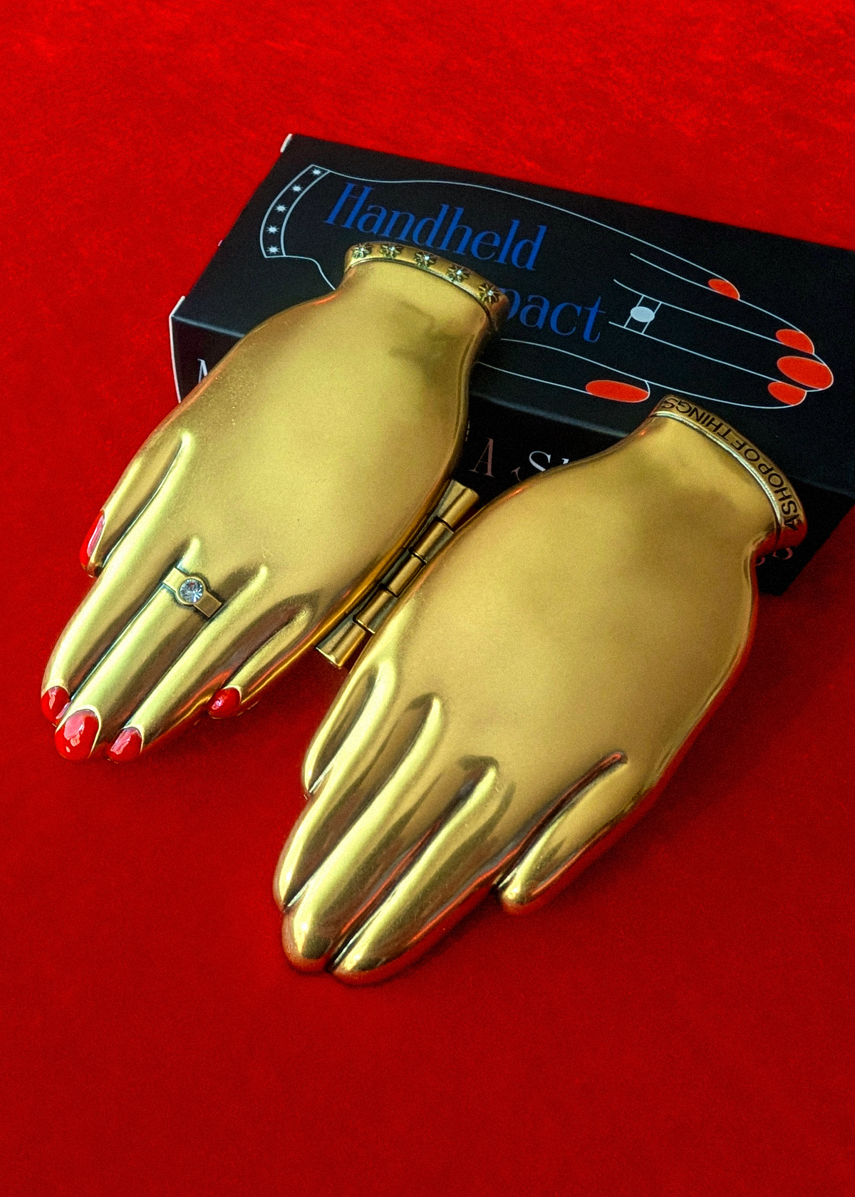 A gold hand-shaped compact mirror with red nail details and a ring on the finger, displayed next to its black packaging box on a red background.