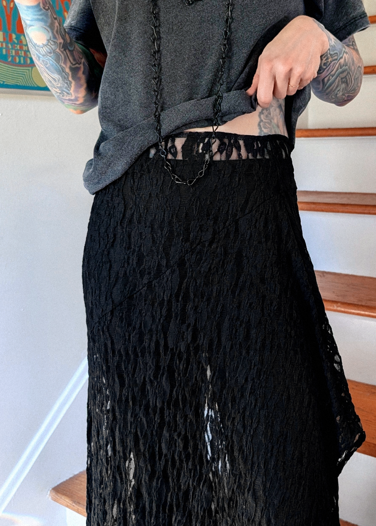 A person wearing a long black lace skirt and a burnout black tee, standing indoors near wooden stairs.  The photo displays the waistline details of the skirt.