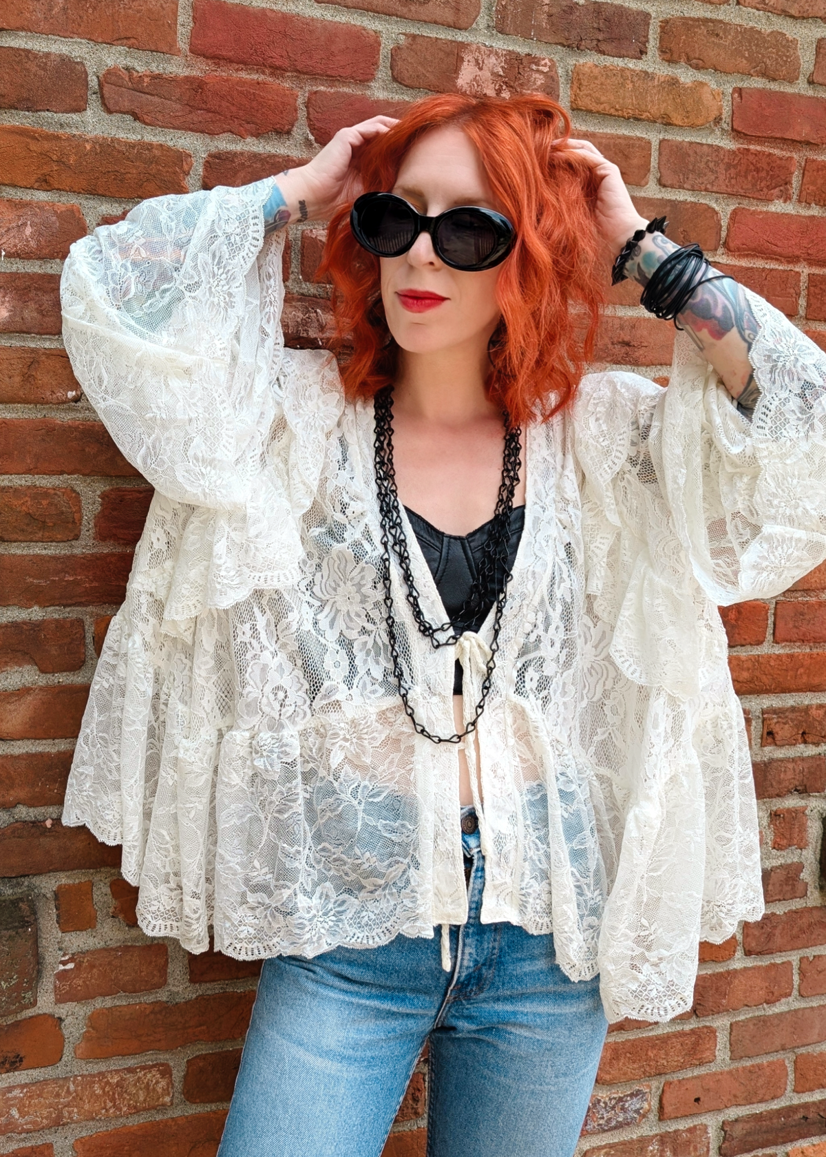 A woman with vibrant red hair and black sunglasses stands against a red brick wall, wearing an ivory lace bohemian-style top over a black corset and blue jeans.
