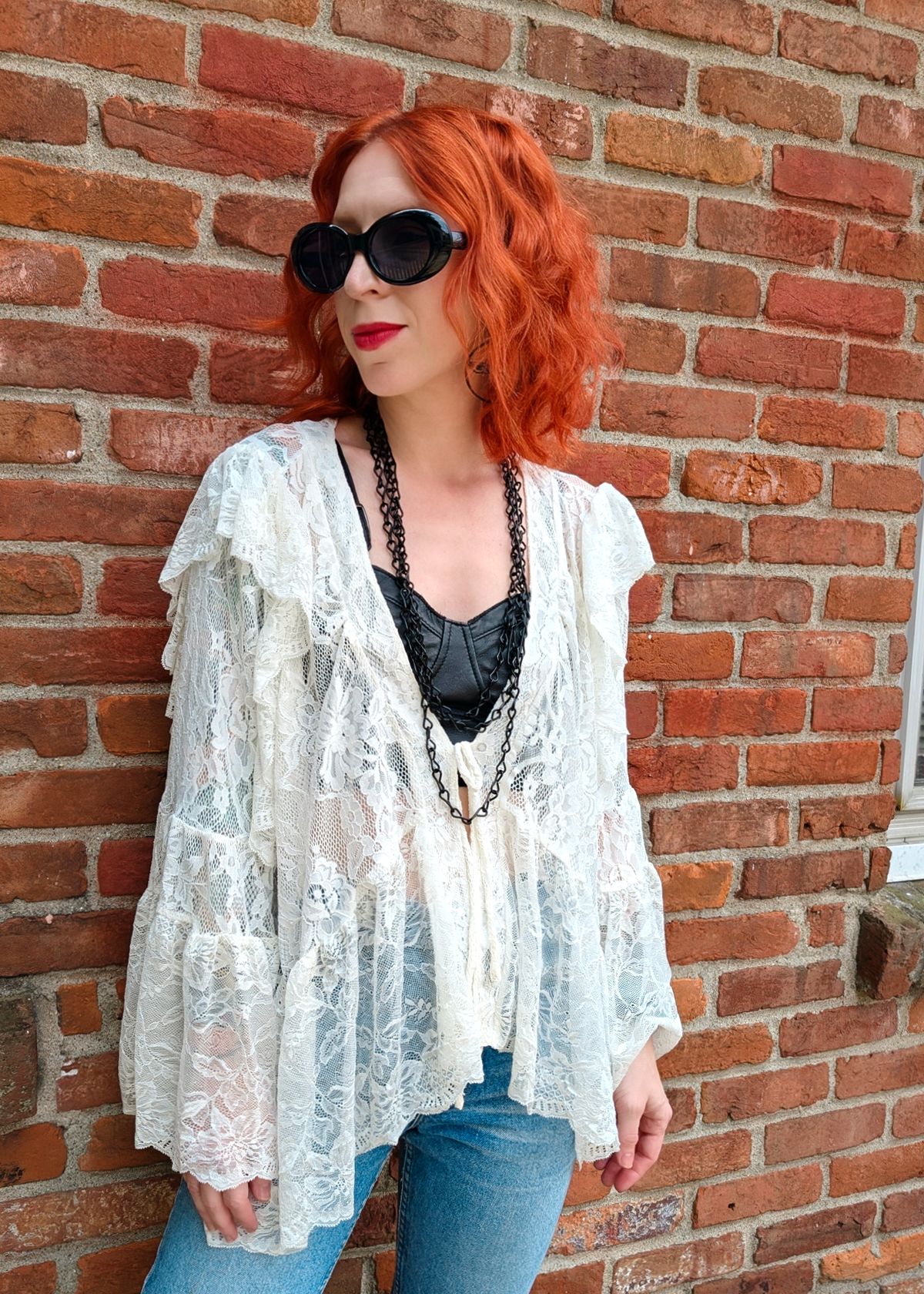 A woman with bright red hair and black sunglasses stands against a red brick wall, wearing a white lace open-front  top over a black lace corset top and blue jeans.