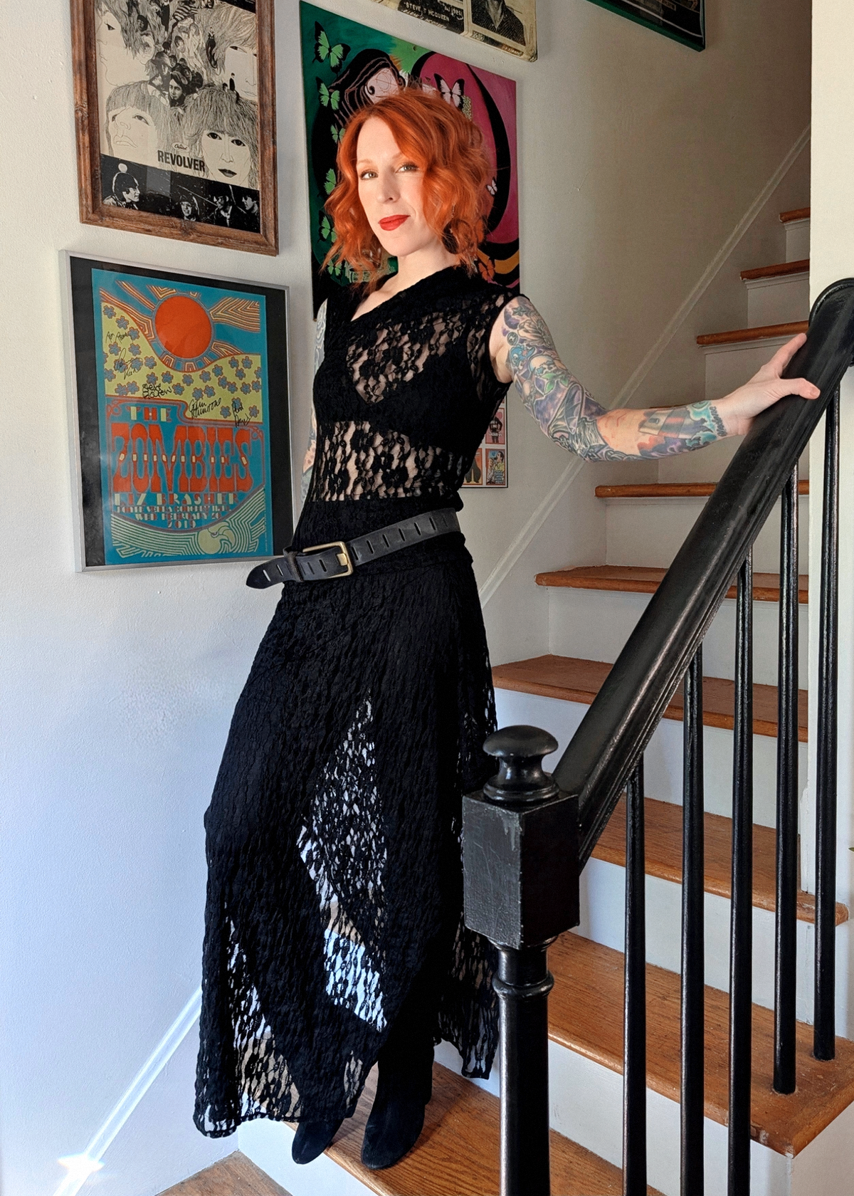 A woman with red hair and tattoos stands on wooden stairs holding a black banister, wearing a black lace top and matching sheer black skirt with black leggings, black tall boots, and a wide black belt slung at the waist