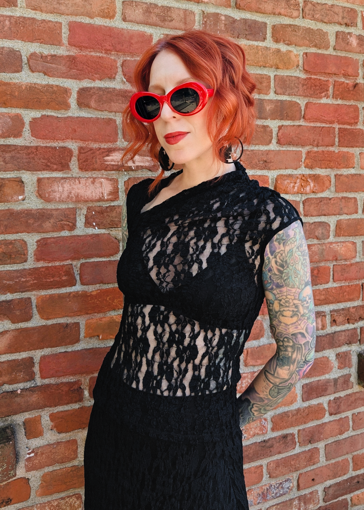 A woman with red hair and tattoos wearing a black lace asymmetrical neck top and matching sheer black lace skirt. Styled with red sunglasses and black earrings, posing against a red brick wall.