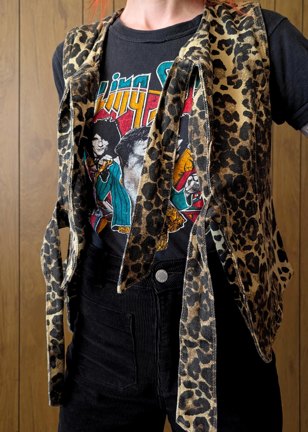 Person wearing a leopard print tie denim vest over a graphic t-shirt against a wooden background