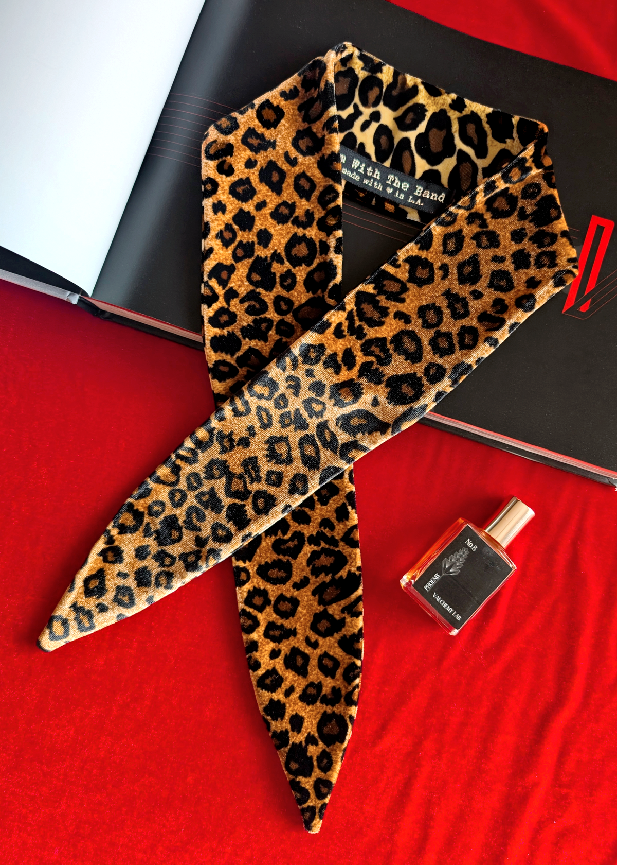 Leopard print velvet scarf tie on a red surface with a black book and perfume bottle.