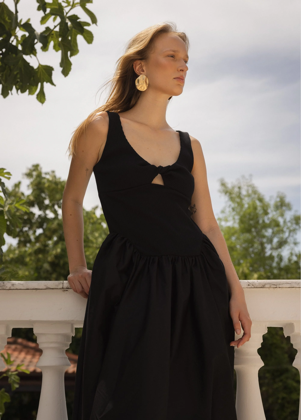 Woman in a black dress standing outdoors with greenery and a white railing in the background