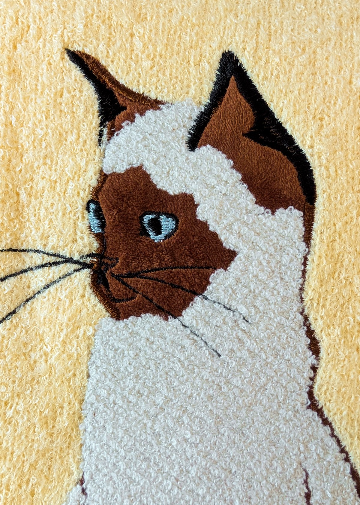 Close-up of a textured sweater featuring a stylized cat portrait with brown and cream fur details on a pale yellow background.
