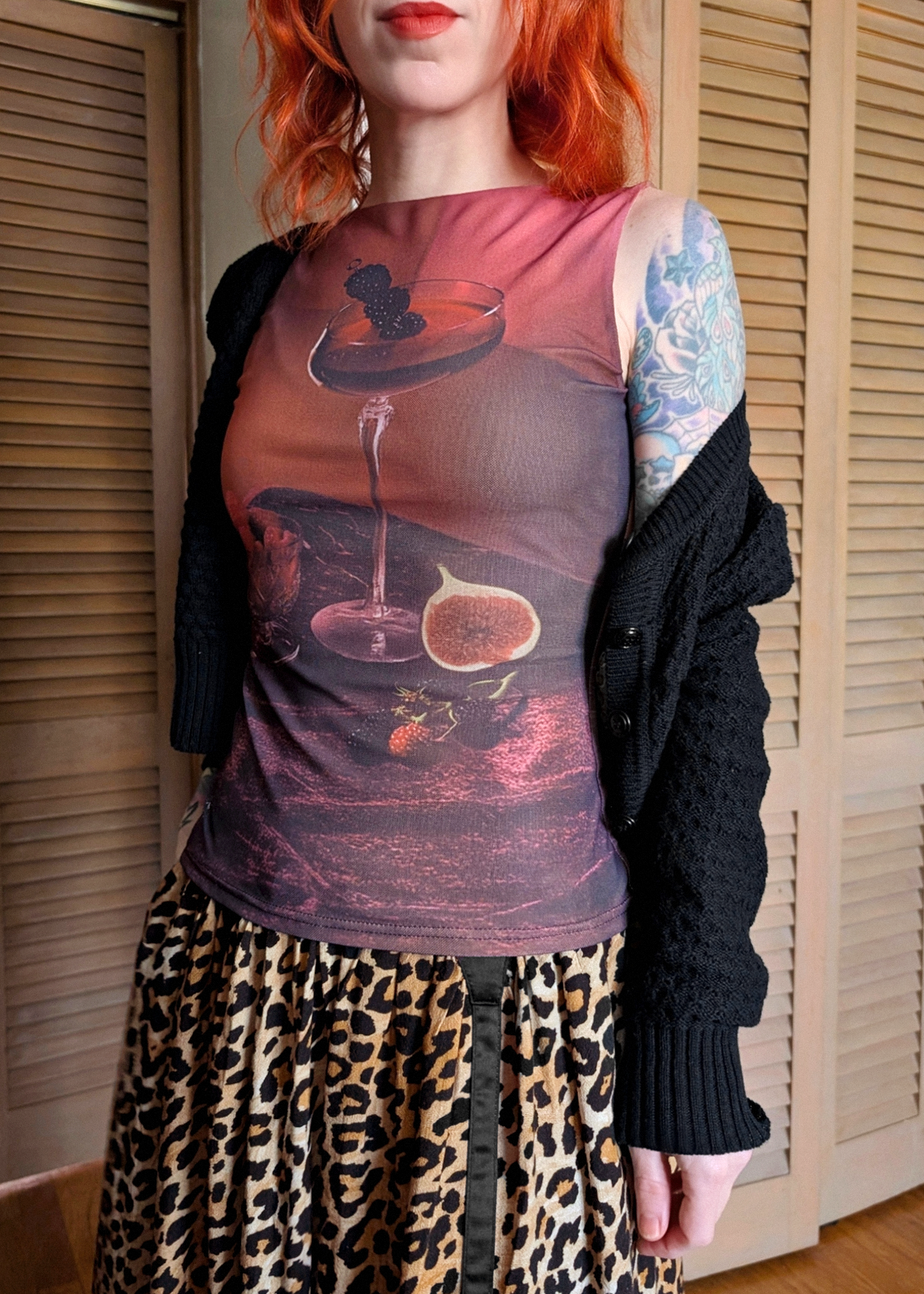 A person wearing a sheer pink and purple graphic top with a printed cocktail and fig still life, styled with an open black cardigan and a leopard-print skirt, standing in front of beige wooden shutters.