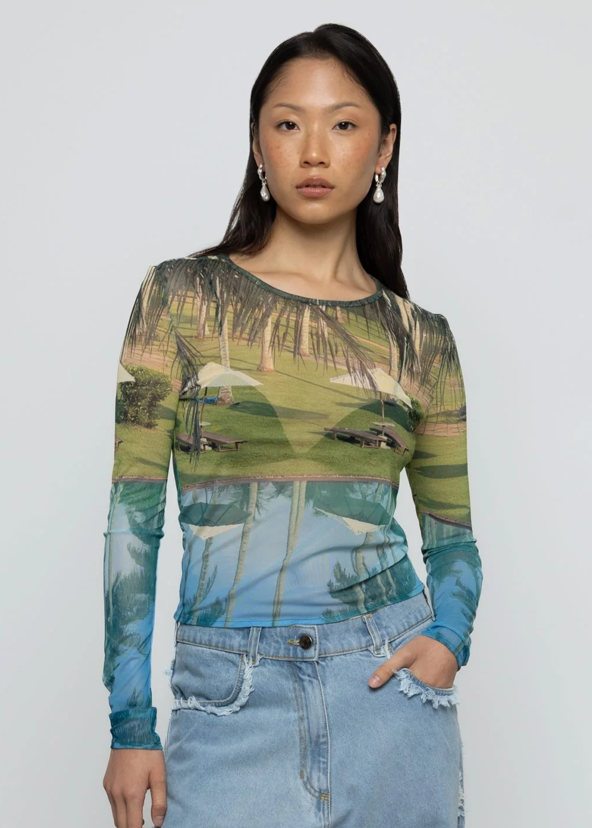 Woman wearing a long-sleeve top with tropical-themed print and blue jeans against a plain background