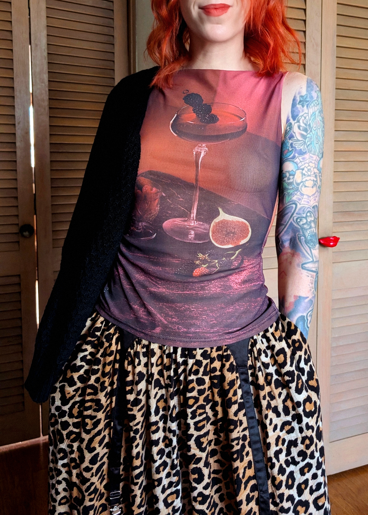 A woman wearing a sleeveless top with a wine glass and fig print, styled with a leopard-print skirt and a black cardigan, standing in front of wooden shutters.
