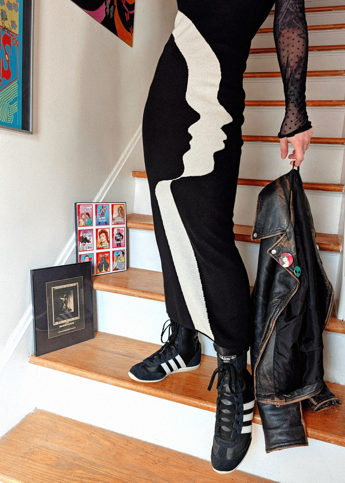 Person wearing a black and white knit dress with Adidas shoes, standing on a staircase holding a jacket.