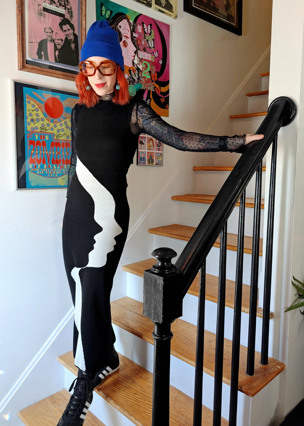 Person in a black and white knit dress with a blue beanie standing on a staircase.