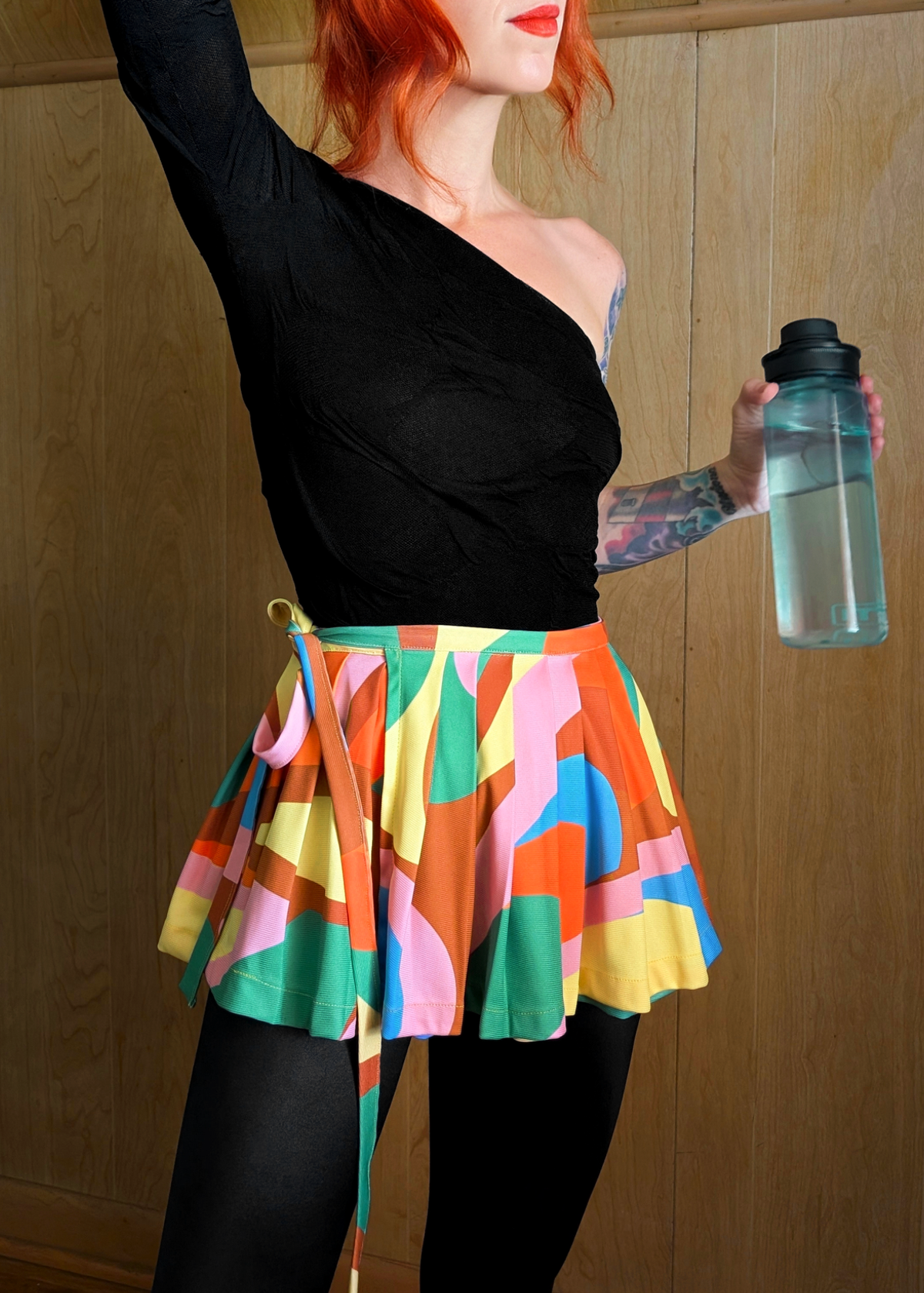 Person wearing a colorful mini belt wrap dance skirt and black top, holding a water bottle against a wooden background