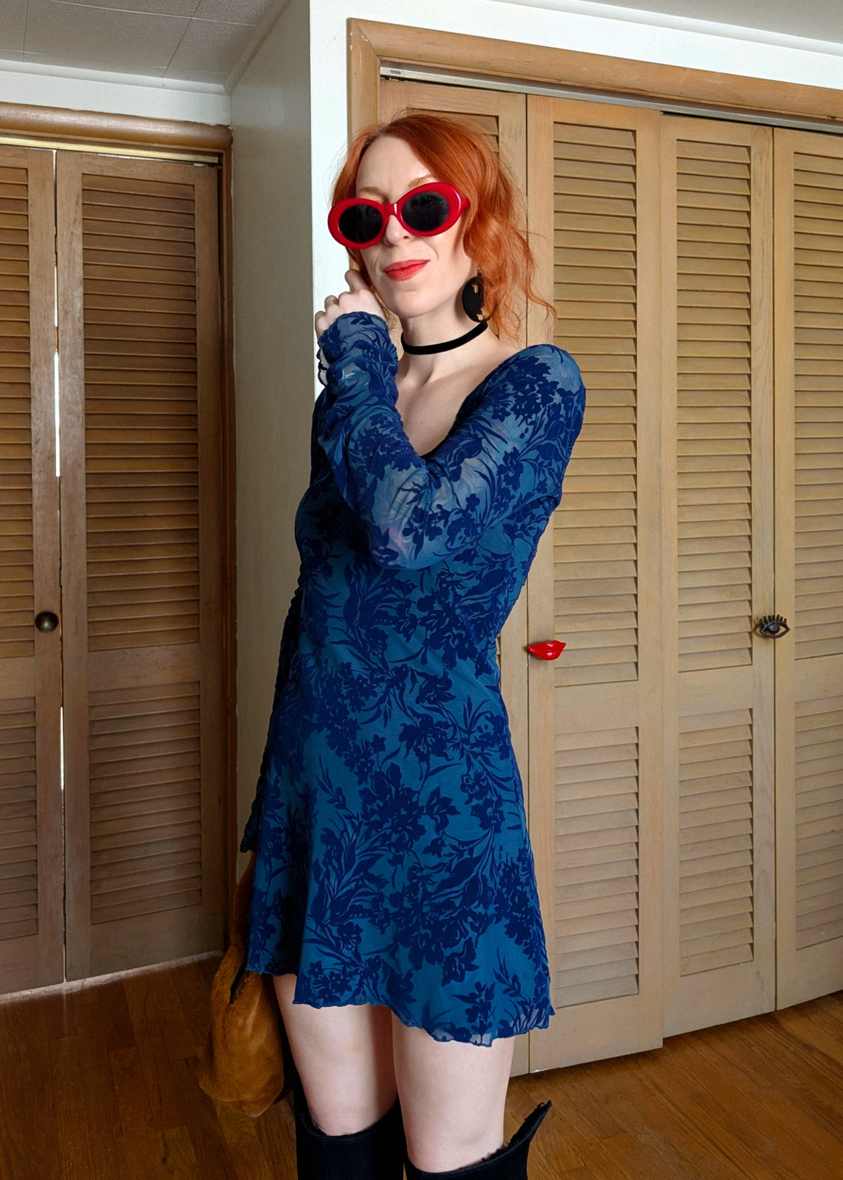 Woman wearing a blue flocked floral mesh dress and red sunglasses in a room with wooden shutters.