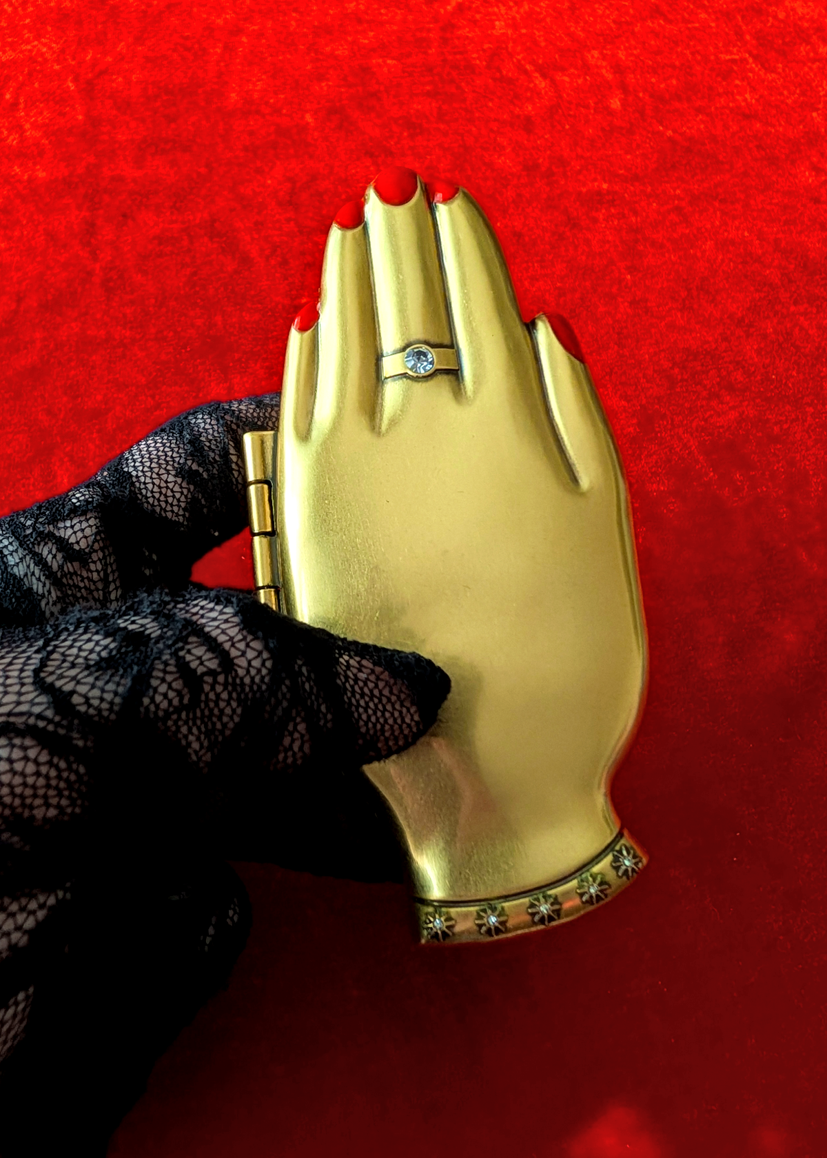 A gold hand-shaped compact mirror with a ring detail on the middle finger, held by a black lace-gloved hand against a textured red background.