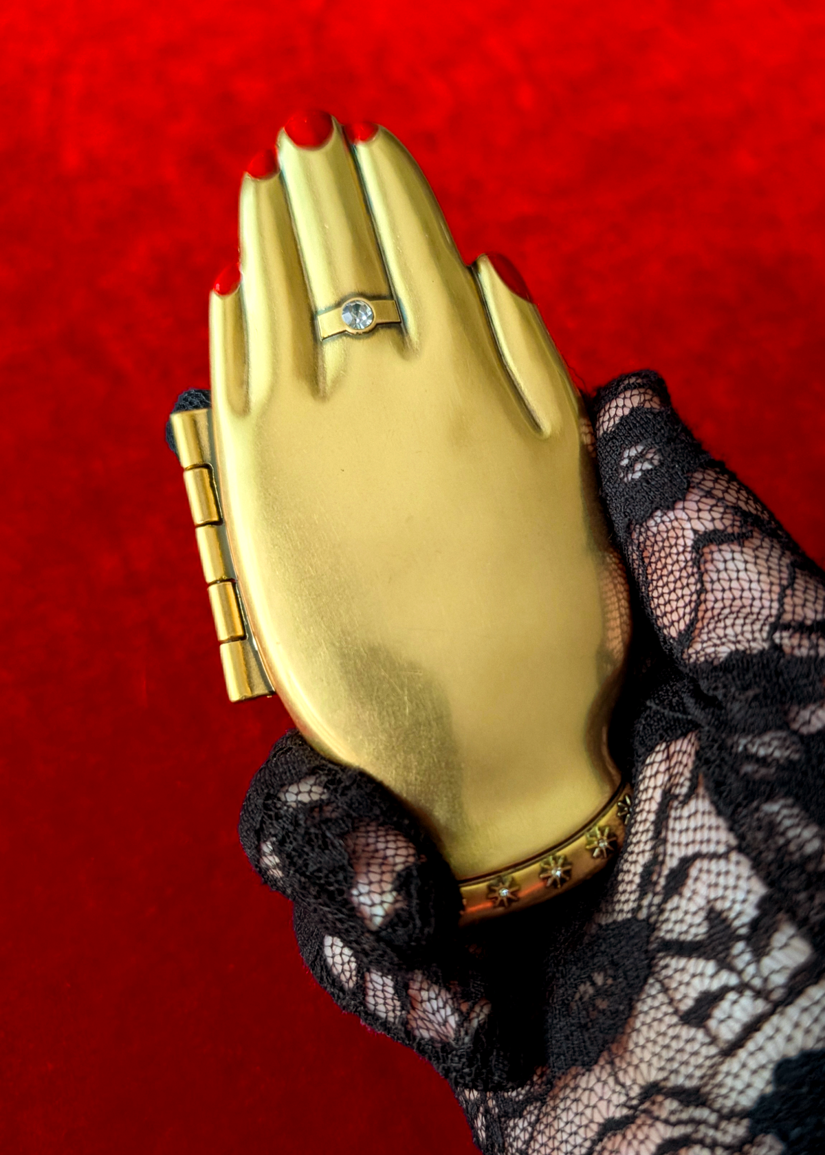 A gold hand-shaped compact mirror with a hinge on the side, held by a person's hand wearing black lace glove against a textured red background. The case features a ring with a clear gemstone on the middle finger