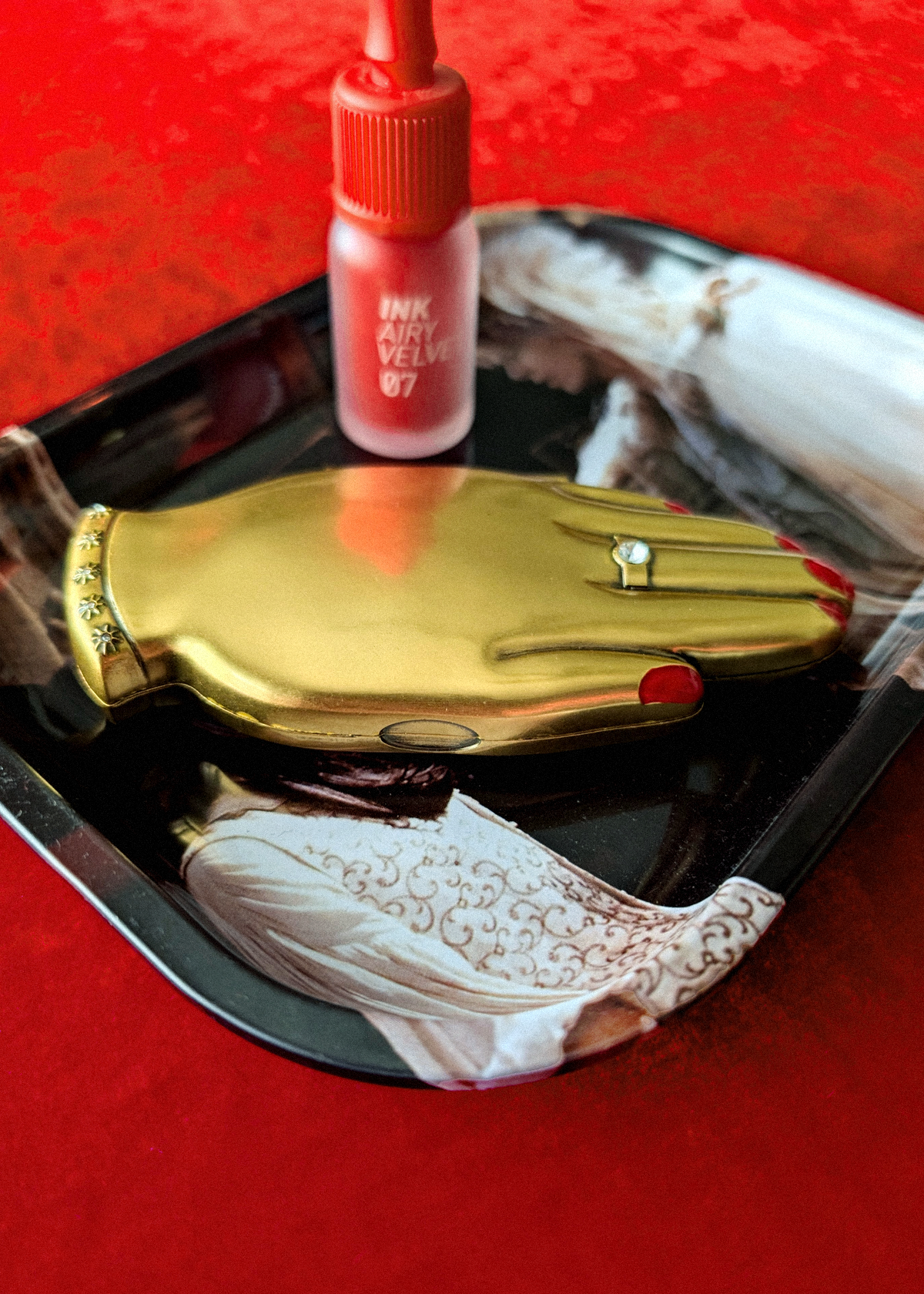 A gold hand-shaped compact mirror with red nail polish details and a small ring, displayed on a black tray with a tube of red lipstick, laying on a Labyrinth movie metal tray