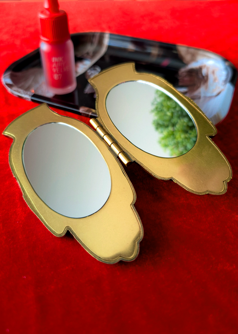 A pair of gold-framed hand-shaped compact mirrors open on a red velvet surface, with a lipstick in the background