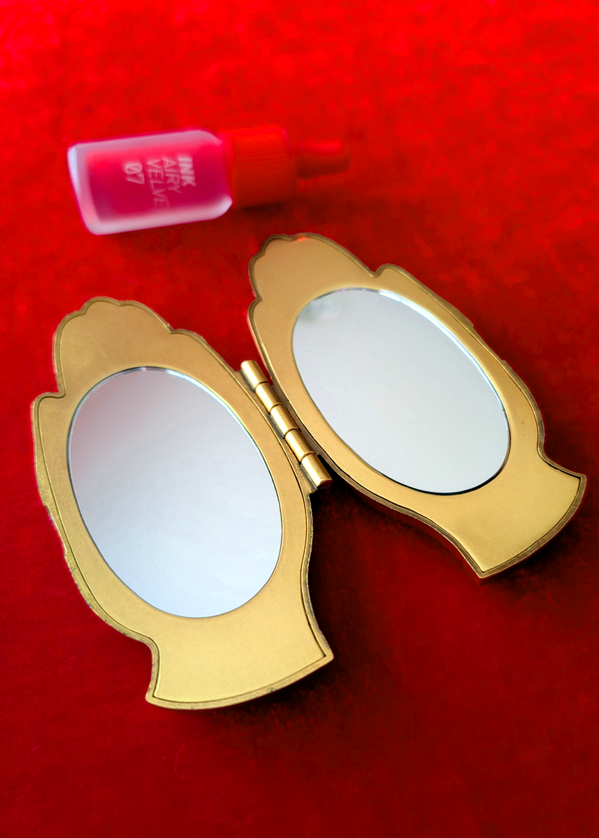 A gold compact mirror with an ornate hand shape lies open on a bright red textured background, with a tube of lipstick in the background