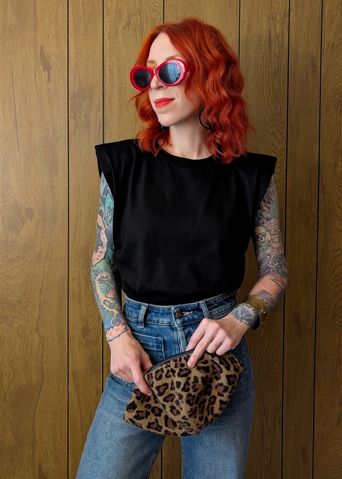 Person with red hair and tattoos wearing a black sleeveless top and blue jeans, holding a leopard print clutch against a wooden background.