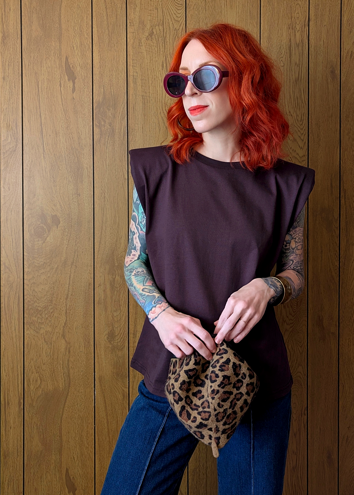 Person with red hair and tattoos wearing sunglasses and a dark plum sleeveless top against a wooden wall.
