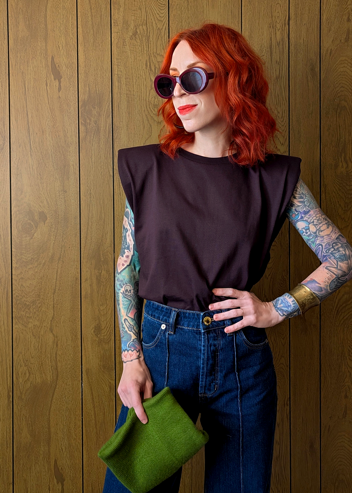 Woman with tattoos wearing a dark plum sleeveless top and blue jeans, holding a green clutch against a wooden background.