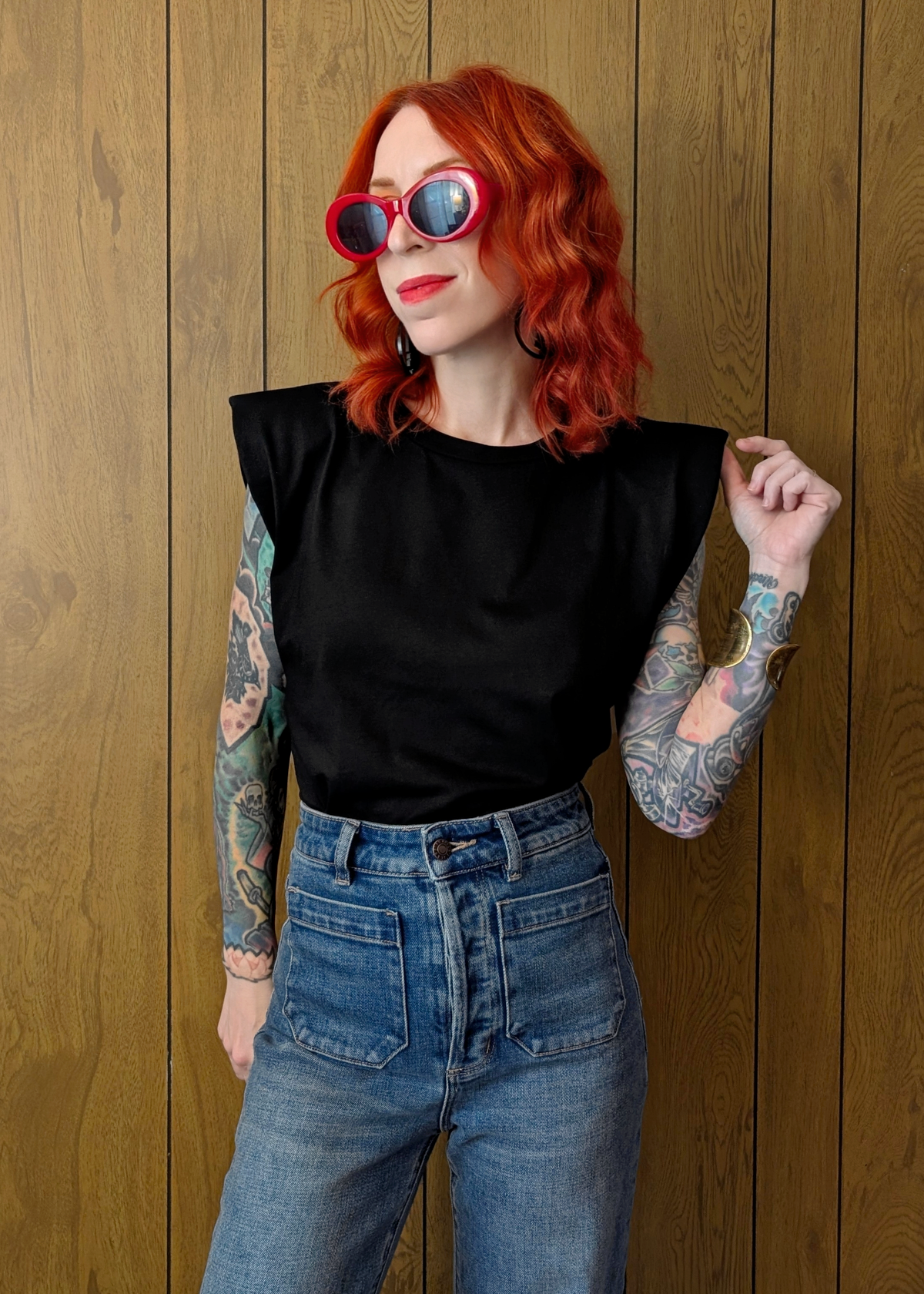 Person with red hair and tattoos wearing a black top and blue jeans against a wooden background