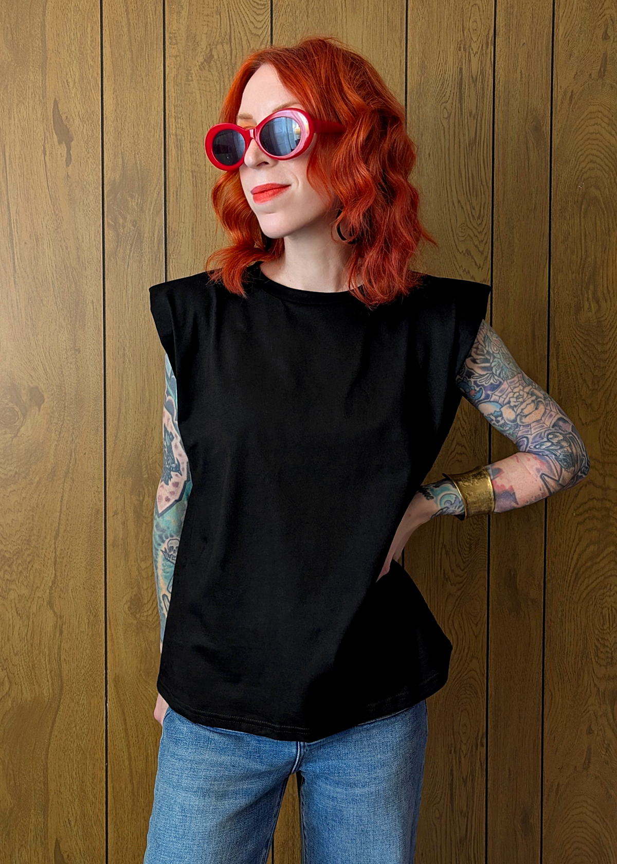 Person with red hair and tattoos wearing a black sleeveless top and blue jeans against a wooden background
