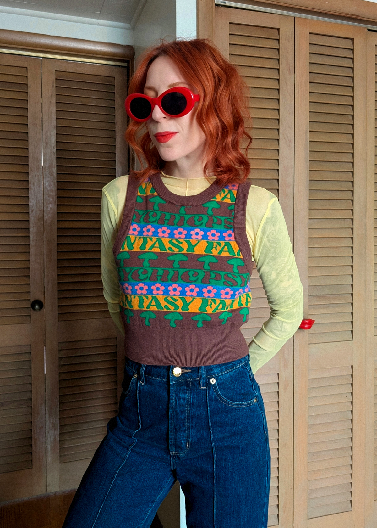 Person wearing a patterned knit vest and blue jeans with red sunglasses indoors.