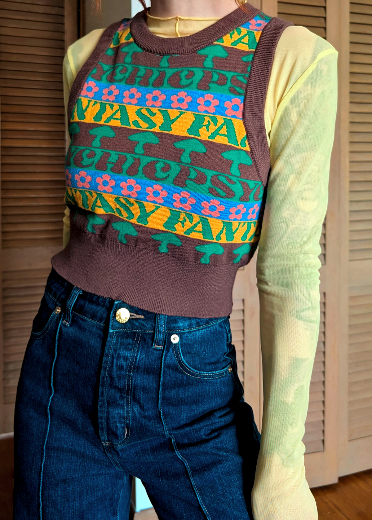Person wearing a colorful patterned sweatervest  over a yellow sheer shirt with blue jeans.