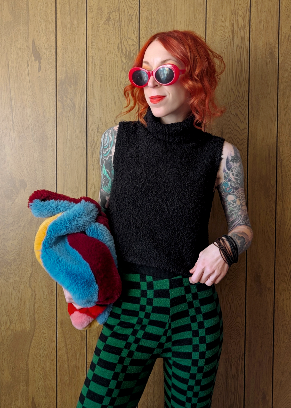 Person with red hair and tattoos wearing a black sleeveless top and green checkered pants, holding colorful socks against a wooden background.